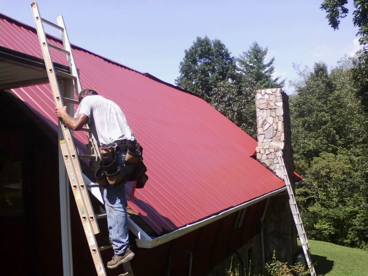 Roofer on a ladder beside a red metal roof and stone chimney — Soffit & Fascia Repair by Roofing Brothers in Youngtown